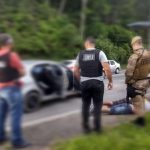 policiais de colete em pé em volta de homens deitados algemados em acostamento. carros em volta; imagem fora de foco