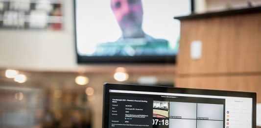 deputados vistos em transmissão em tela de notebook e outro deputado ao fundo em telão