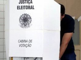 homem atrás de cabine de votação com o símbolo da justiça eleitoral