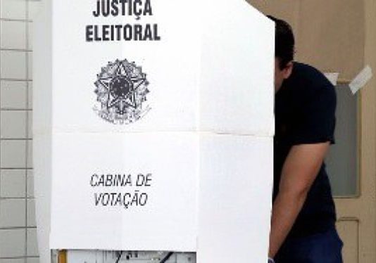 Desembargador destaca ocorrências e apresenta cenário das eleições em SC homem atrás de cabine de votação com o símbolo da justiça eleitoral