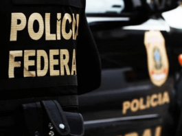 Obstrução: Uma pessoa é presa em nova fase da Operação Alcatraz agente visto de costas sentado em uma bancada com laptop usando colete escrito polícia federal e com uma viatura na sua frente