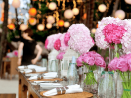 Evento com flores brancas e rosas em vasos de vidro em cima de uma mesa de madeira; guardanapos brancos com talheres sob a mesa também; fundo com luzes desfocadas