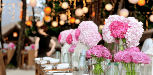 Volta de eventos e turismo em SC será tema de audiência pública Evento com flores brancas e rosas em vasos de vidro em cima de uma mesa de madeira; guardanapos brancos com talheres sob a mesa também; fundo com luzes desfocadas