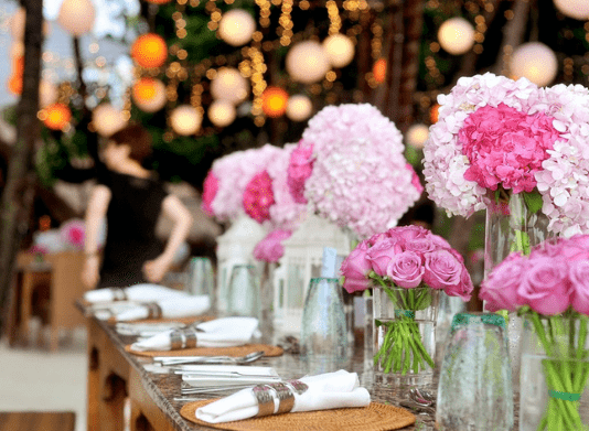 Evento com flores brancas e rosas em vasos de vidro em cima de uma mesa de madeira; guardanapos brancos com talheres sob a mesa também; fundo com luzes desfocadas