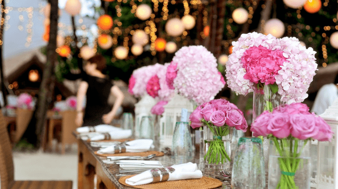 Evento com flores brancas e rosas em vasos de vidro em cima de uma mesa de madeira; guardanapos brancos com talheres sob a mesa também; fundo com luzes desfocadas