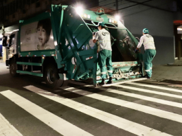 Florianópolis muda horários da coleta de lixo no Continente Dois homens na parte de trás de um caminhão de lixo verde em Florianópolis