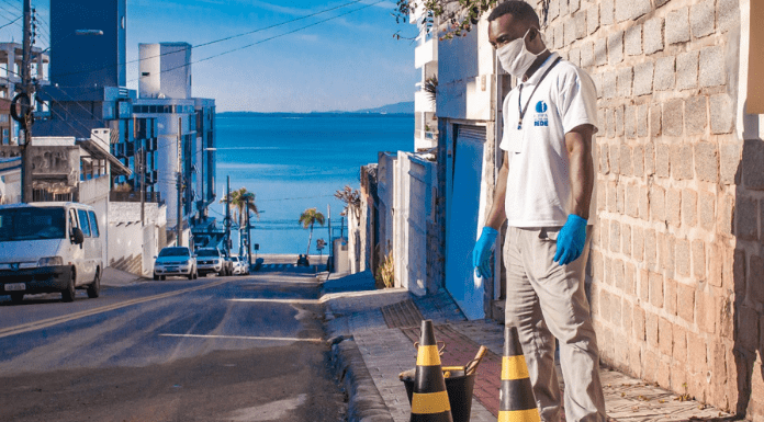 Fiscal parado em pé perto do esgoto do lado direito, está usando máscara branca e luvas azuis; o restante da foto é composto por casas, apartamentos, carros e no final uma praia da capital