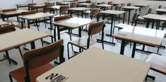 Mesas e cadeiras de sala de aula vazias; na mesa há a logo do Governo do Estado de Santa Catarina