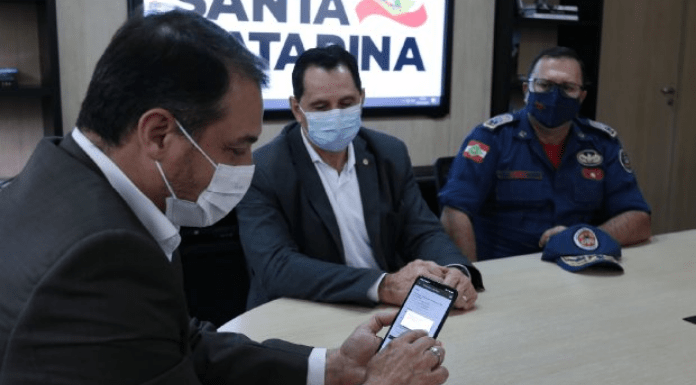 O governador Carlos Moisés, com máscara, sentado a esquerda e mexendo no celular; no resto da extensão da mesa mais dois homens sentados, com máscara e olhando para o governador do Estado