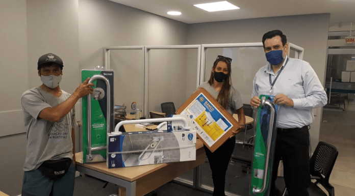Um homem a esquerda de camisa e máscara cinza, segurando material doado pela cooperativa; no centro uma mesa com alguns materiais; e a direita uma mulher segurando uma caixa fechada e embalada, e um homem segurando um material de hidraulica
