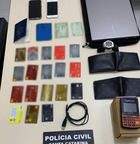Polícia Civil prende dois homens por estelionato em Florianópolis Cartões de crédito clonados, carteiras, celulares e uma placa da Polícia Civil de Santa Catarina em cima da mesa