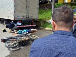PRF simula acidentes graves de moto para conscientizar motociclistas para conscientizar motociclistas: homem olha moto e boneco caídos atrás de caminhão, com prf ao lado