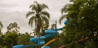 Parque aquático: árvores ao redor do tobogã azul, da piscina com um homem dentro e de cadeiras brancas que estão na borda da piscina