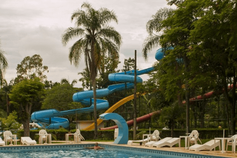 Parque aquático: árvores ao redor do tobogã azul, da piscina com um homem dentro e de cadeiras brancas que estão na borda da piscina