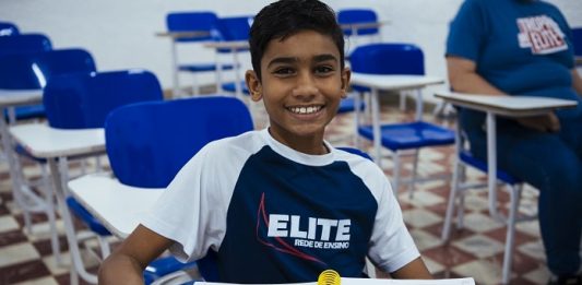 Colégio Elite abre inscrições para prova de bolsas Menino em sala de aula do Elite Rede de Ensino, com uniforme da instituição e com o caderno aberto em cima da mesa