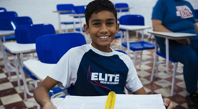 Menino em sala de aula do Elite Rede de Ensino, com uniforme da instituição e com o caderno aberto em cima da mesa