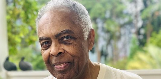 gilberto gil é congratulado com título de cidadão honorário; ele olha para foto; usa camisa com foto de criança