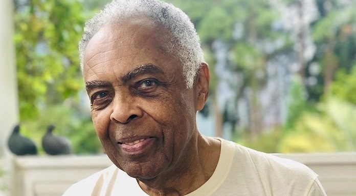 gilberto gil é congratulado com título de cidadão honorário; ele olha para foto; usa camisa com foto de criança