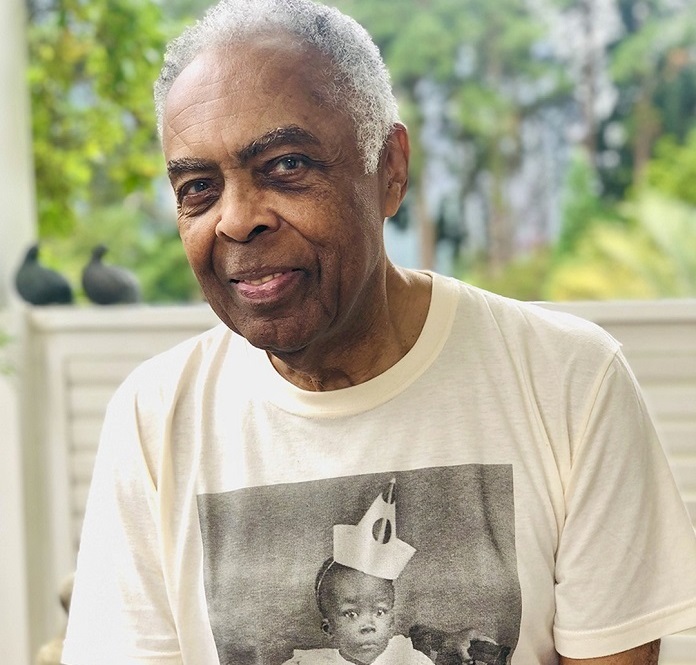 gilberto gil é congratulado com título de cidadão honorário; ele olha para foto; usa camisa com foto de criança