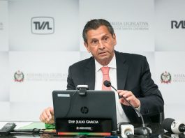 em entrevista de final de presidência da alesc, julio garcia está à frente de um note book com microfone; ao fundo um painel de logos da alesc