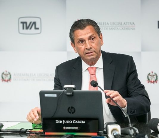 em entrevista de final de presidência da alesc, julio garcia está à frente de um note book com microfone; ao fundo um painel de logos da alesc