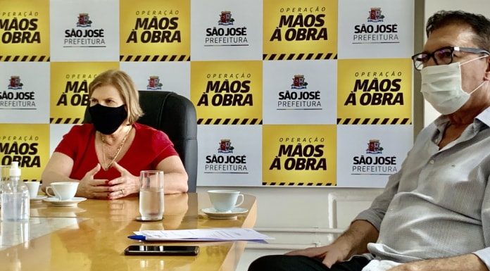 são josé transição governo na ponta de uma grande mesa adeliana fala gesticulando; orvino sentado ao lado de perna cruzada; ambos de máscara; copos de água sobre a mesa e painel da prefeitura ao fundo