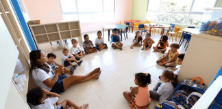Uma professora e 14 alunos pequenos sentados no chão de uma sala de aula