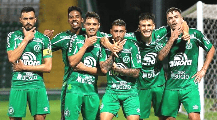 Seis jogadores da Chapecoense com uniforme verde principal, parados em fileira, fazendo pose após gol marcado pela equipe