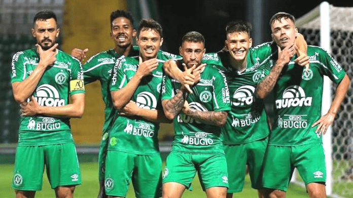 Seis jogadores da Chapecoense com uniforme verde principal, parados em fileira, fazendo pose após gol marcado pela equipe