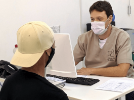 Homem com uniforme da Prefeitura de Biguaçu, sentado de um lado da mesa e mexendo no computador, e um segundo homem, paciente, de costas esperando ser marcada a sua consulta - Consultas de rotina voltam a ser agendadas nos postos de saúde de Biguaçu