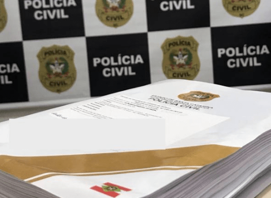 Inquérito policial finalizado em cima de uma mesa na delegacia, atrás o painel da Polícia Civil de Santa Catarina