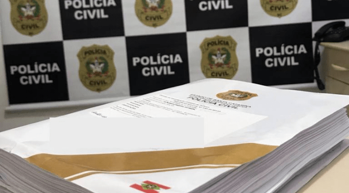 Inquérito policial finalizado em cima de uma mesa na delegacia, atrás o painel da Polícia Civil de Santa Catarina