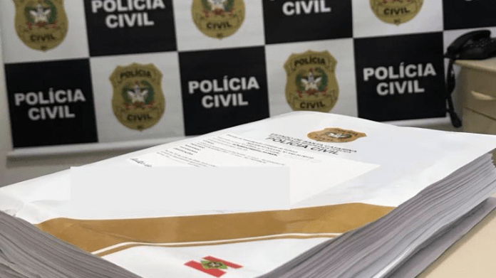 Inquérito policial finalizado em cima de uma mesa na delegacia, atrás o painel da Polícia Civil de Santa Catarina