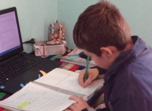 Criança em casa, com notebook em sua frente e escrevendo em um caderno
