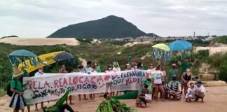 Moradores do Santinho protestam com faixa "pela realocação das elevatórias de esgoto"; alguns vestidos de jacaré; morro das aranhas e dunas ao fundo
