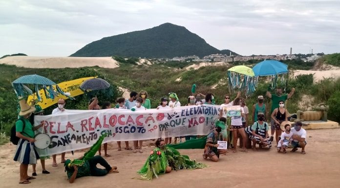 Moradores do Santinho protestam com faixa "pela realocação das elevatórias de esgoto"; alguns vestidos de jacaré; morro das aranhas e dunas ao fundo