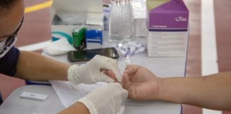 profissional de saúde usando luvas faz teste rápido em dedo de pessoa com a mão sobre mesa - São José tem 462 casos ativos