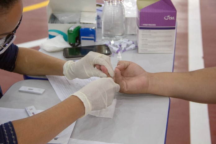 profissional de saúde usando luvas faz teste rápido em dedo de pessoa com a mão sobre mesa - São José tem 462 casos ativos