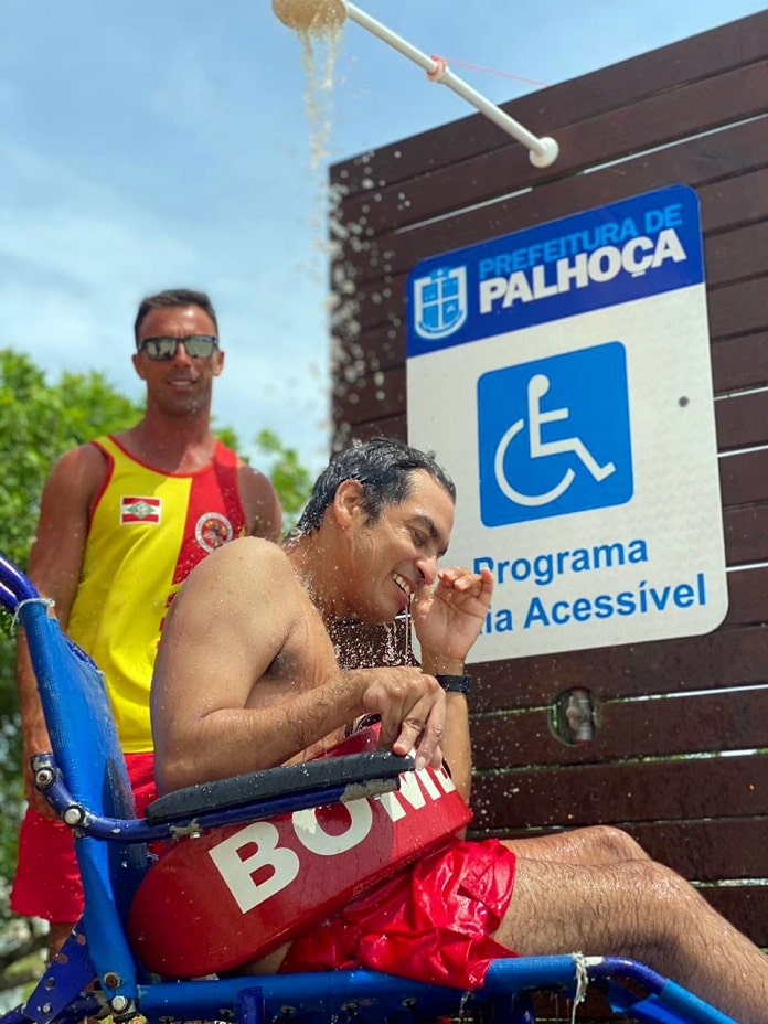 dentro do programa de acessibilidade nas praias de palhoça, cadeirante toma banho em chuveiro externo em cadeira de rodas especial observado por guarda-vidas