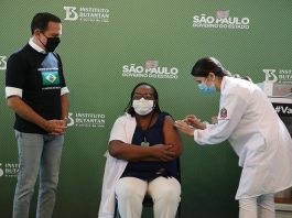 monica, primeira vacinada contra coronavírus, sentada recebe a dose no braço dada pela enfermeira, observadas pelo governador de são paulo, joão dória, todos usam máscaras