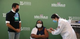 monica, primeira vacinada contra coronavírus, sentada recebe a dose no braço dada pela enfermeira, observadas pelo governador de são paulo, joão dória, todos usam máscaras