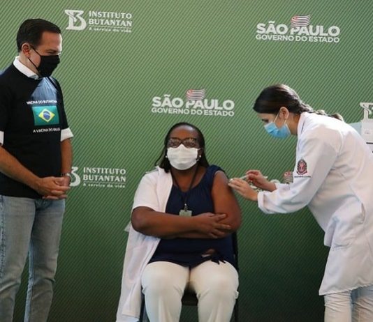 monica, primeira vacinada contra coronavírus, sentada recebe a dose no braço dada pela enfermeira, observadas pelo governador de são paulo, joão dória, todos usam máscaras