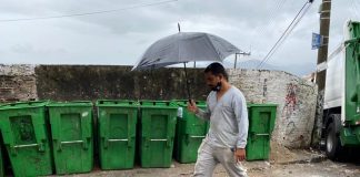 para ilustrar roteiro de coleta de lixo em florianópolis, homem com guarda chuva passa em frente à contentores de lixo vazios enfileirados