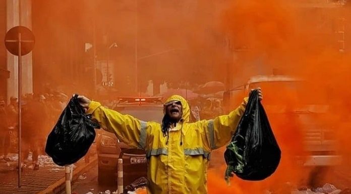 para ilustrar a matéria da decisão da justiça que a comcap deve voltar ao trabalho, foto mostra um funcionário com capa de chuva e braços abertos segurando dois sacos de lixo em meio a grande quantidade de lixo espalhada na rua e fumaça laranja atrás