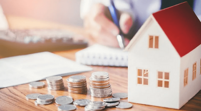 Crédito Imobiliário da CAIXA em 2020: Pessoa no fundo escrevendo em um caderno, no canto direito na frente uma casinha e do lado várias moedas empilhadas