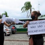 em protesto de greve, mulher simula aplicação de vacina no braço de outra mulher; ela usa uma agulha gigante de cartolina; a outra segura um cartaz escrito "nossas vidas não são descartáveis"; trânsito de são josé ao fundo