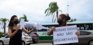 em protesto de greve, mulher simula aplicação de vacina no braço de outra mulher; ela usa uma agulha gigante de cartolina; a outra segura um cartaz escrito "nossas vidas não são descartáveis"; trânsito de são josé ao fundo