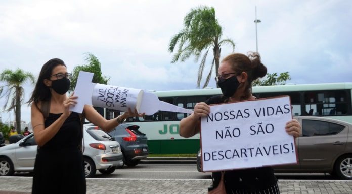 em protesto de greve, mulher simula aplicação de vacina no braço de outra mulher; ela usa uma agulha gigante de cartolina; a outra segura um cartaz escrito "nossas vidas não são descartáveis"; trânsito de são josé ao fundo