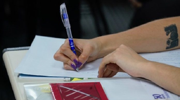 Mão com unhas pintadas de roxo segurando uma caneta de tubo transparente com detalhes em azul, ao lado um bloco vermelho em cima de uma pasta branca. A imagem representa um estudante, o governo de SC anunciou bolsas para ensino superior