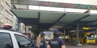 Duas guardas municipais uniformizadas e de costas ao lado de um carro da Guarda Municipal, elas estavam no terminal urbano de Florianópolis, reforçando a fiscalização das medidas do novo decreto.